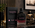 A bottle of Valentino Uomo Born in Roma Intense Eau de Parfum stands next to its black packaging box on a polished dark marble surface. The background is a dimly lit, stylish room with a lamp, glasses, and a watch.