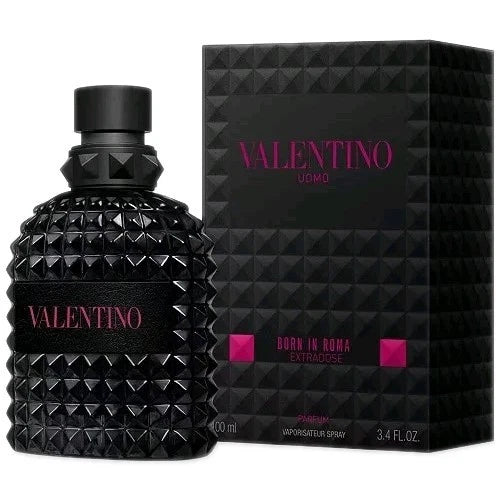 A bottle of Valentino Uomo Born in Roma Intense Eau de Parfum stands next to its black packaging box on a polished dark marble surface. The background is a dimly lit, stylish room with a lamp, glasses, and a watch.